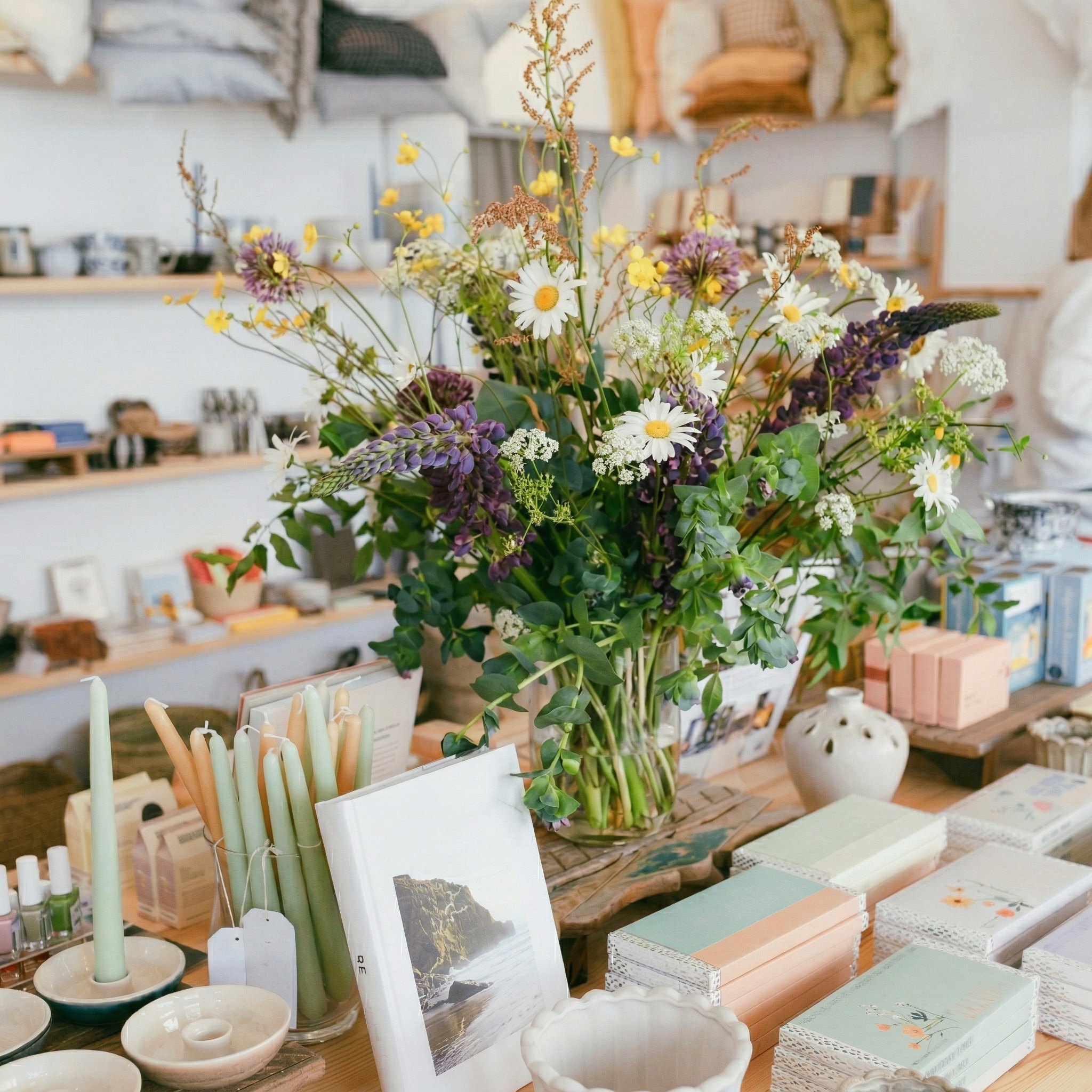 A gift shop table with a jug of wildflowers among cards and candles